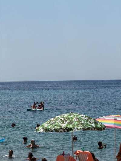 The image depicts a bright, sunny day at a beach in Sithonia, Greece. The scene is divided, with the left side showcasing a clear, pale blue sky and the right side filled with the azure waters of the sea. Several people are engaged in water activities. In the foreground, a colorful beach umbrella with rainbow stripes and another umbrella covered with a green leaf pattern are visible.
On the water, a group of individuals are paddleboarding. One paddleboard has three people on it, with two in the back and one in the front, appearing to be paddling. Another paddleboard is further to the right, with at least one person visible. Scattered throughout the water are other individuals swimming or wading. Some people are looking towards the camera, while others are focused on their activities. The water is calm and reflects the sunlight, creating a shimmering effect. The overall atmosphere suggests a leisurely and enjoyable day at the beach, with people taking advantage of the warm weather and clear water. There is no visible text in the image.