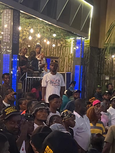 The image depicts an outdoor gathering at night in Oba, Nigeria. A crowd of people, predominantly of African descent, are visible. The scene is illuminated by decorative string lights and blue LED lights integrated into a patterned pillar. In the foreground, several individuals are looking in different directions, with some wearing caps and headscarves. A man in the center foreground wears a white t-shirt with "ADVENTURES" printed on it. The background features a textured wall, possibly made of wood or stone, and foliage. A metal railing is visible, suggesting a secured area or a queue. The overall atmosphere appears to be that of a social event or celebration.