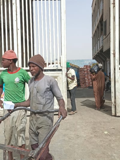 The image depicts an outdoor commercial or industrial scene in Fagge C, Nigeria, under a hazy sky. In the foreground, two males stand on a concrete surface next to a large, partially open white metal gate with vertical bars. The male on the left, possibly an adolescent, wears a red knit cap, a green t-shirt with a "Fagge C" logo, and light-colored cargo shorts. He holds a small white object in his right hand and looks to the left. Adjacent to him, a bearded adult male wears a brown knit cap, a dirty light grey short-sleeved tunic, and dark trousers. He holds the metal handlebars of a rudimentary metal handcart or trolley and looks towards the right.
Through the open gate, in the mid-ground, other individuals and stacked goods are visible. To the left, a male in a green knit cap, light yellow shirt, and dark trousers leans against a structure, looking right. To the right, another male, dressed in a brown robe, carries a black object on his head and a white string in his left hand, walking away from the gate opening. Behind these individuals, numerous stacks of red-orange rectangular objects, likely packaged goods or building materials, are visible, appearing to be loaded onto a large vehicle or trailer. The right side of the frame is dominated by a multi-story, light brown concrete building with balconies or windows. The ground is a light-colored concrete surface, with scattered debris visible.
