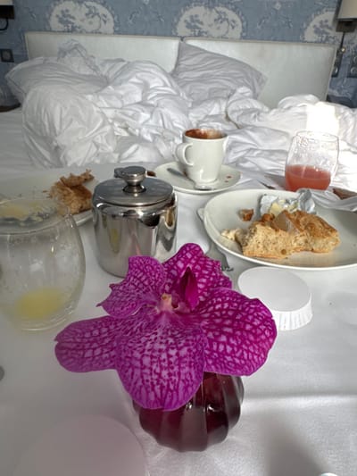 The scene depicts a recently concluded breakfast in a hotel room in Deauville, France. In the background, a large bed is visible with a rumpled white duvet and pillows, situated against a white upholstered headboard. The wall behind features blue and white patterned wallpaper, with two wall-mounted light fixtures partially visible.
In the foreground, on a white surface, various items associated with breakfast are arranged. A vibrant magenta orchid with dark speckles stands in a small, dark red fluted glass vase at the center-right. To its left, a silver-colored, lidded pot is positioned near an empty clear glass containing a small amount of yellow liquid residue. A white ceramic coffee cup with a handle, holding dark liquid residue, rests on a matching saucer with a spoon. Adjacent, a clear glass contains a partial volume of reddish-pink liquid. Two white oval plates are present: one holds several pieces of bread, butter in a foil wrapper, and a partially visible fork; the other contains bread crumbs and yellow residue. A small, white fluted paper cup or container is also visible on the right. No individuals are present in the image.