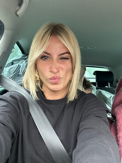 The image displays a blonde Caucasian female secured by a grey seatbelt inside a vehicle, located in Brignoles, France. She occupies the foreground, facing the camera with a playful expression: her left eye is closed in a wink, her lips are pursed, and her nose is lightly scrunched. She wears a black long-sleeved top and two distinctive gold-colored, abstract-shaped earrings. In the background, partially obscured, a second individual with dark, curly hair is visible, seemingly resting in a rear passenger seat. A striped blue and white bag or textile is present in the rear left, and a reddish, textured fabric is partially visible on the far right. The car's interior features light grey overhead upholstery and black headrests. The external view through the rear window is blurred, indicating an overcast or dim daytime environment.