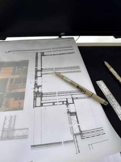 The scene depicts a desk setup located in Newcastle upon Tyne, United Kingdom, centered on architectural documentation. A white sheet of paper, displaying technical architectural drawings, occupies the midground. The primary drawing is a detailed cross-section illustrating building wall and floor construction elements, rendered in black lines and hatching. To the left, a blurred photographic image of a modern building with wooden cladding is visible underneath or alongside the main drawing.
A beige-bodied technical drawing pen, identified as a Pigma Micron 005, rests horizontally across the architectural drawing. To the right of the paper, on a dark, flat surface, a second identical pen is partially visible along with a metallic ruler, approximately 15 cm (6 inches) in length, showing both metric and imperial markings. The background features the bottom bezel of a black computer monitor, with the "ADX" brand logo visible. The monitor screen is dark. No individuals are present. The arrangement suggests an active architectural design or study environment.