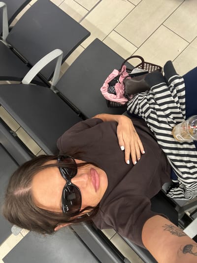 A woman wearing a brown t-shirt and black sunglasses is lying on a row of black chairs. She has white nail polish and a tattoo on her arm. A striped sweater, a plastic cup with a drink, and a bag are on the chair next to her. The scene appears to be indoors, likely at an airport. The location may be in Crawley, United Kingdom.