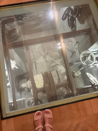 The image shows a view through a glass floor into an archaeological excavation site in Gatchina, Russia. The site appears to be an old industrial or workshop area, with remnants of machinery, stone structures, and wooden beams visible. In the foreground, a pair of bare feet wearing beige sandals are visible, standing on a wooden floor. The lighting in the excavation site is dim, with some artificial light sources casting shadows. The glass floor has some reflections and smudges, which partially obscure the view of the site below. The overall impression is one of historical preservation and public accessibility to an archaeological find.