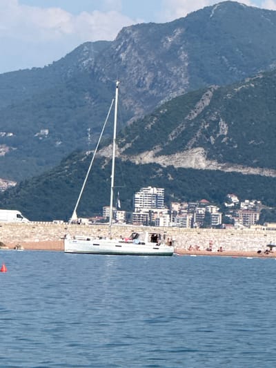 The image captures a daytime scene of the Adriatic Sea with a coastal town and mountainous backdrop, likely Sveti Stefan, Montenegro. In the foreground, the calm, blue water is partially visible, with a bright orange buoy floating to the left. A white sailboat with a tall mast is moored near the shore, with several people visible on its deck.

The middle ground features a stone-walled embankment, behind which a beach with scattered sunbathers is visible. Beyond the beach, a cluster of buildings, including modern high-rise structures and smaller residential buildings, ascend the hillside. The mountains in the background are covered in dense green vegetation and show rocky outcrops and some areas of exposed earth. The sky is partly cloudy. The scene suggests a warm, sunny day at a popular coastal resort.