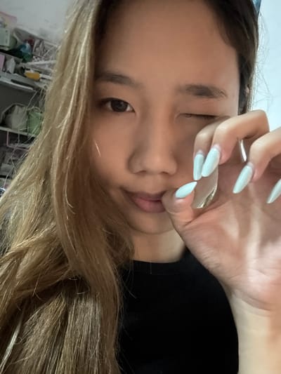A young Asian woman with long, blonde hair is in a close-up shot, appearing to be indoors. She is winking with her left eye, while her right eye is open. Her right hand is raised to her face, with her fingers touching her lips. She has long, almond-shaped fingernails painted a light blue color. She is wearing a black top. The background appears to be a shelf with various items, suggesting a cluttered environment. The image is likely taken in Nan'an District, China.