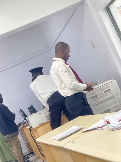 The image depicts an indoor office or administrative area in Zaria, Nigeria. Three individuals are present within a brightly lit space featuring white walls and a light-colored ceiling with visible support beams.
In the mid-ground, a male subject with short hair, wearing a white long-sleeve shirt, a dark red tie, and dark trousers, stands with his back partially to the camera, leaning towards a large, light-colored multifunction copier machine. He holds a red pen or marker in his right hand near the copier's control panel. Directly behind him, another male subject, also in a white long-sleeve shirt and dark trousers, wears a dark peaked cap. He is partially obscured and appears to be standing at a workstation with a computer monitor. Further to the left, a third individual, mostly visible from the waist down, wears a dark jacket, a light green garment, and light-colored trousers, standing at another workstation with a computer monitor.
The foreground features a light-colored desk or counter with stacks of white papers and a clear plastic holder containing additional papers, with the partial letters "UBE" visible on it. Multiple computer monitors and other office equipment are visible on desks throughout the scene. A white electrical outlet is mounted on the wall to the far right, adjacent to a visible window frame.