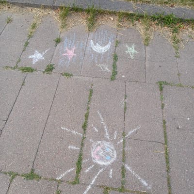 The image shows a section of sidewalk in Baranavichy, Belarus, featuring children's chalk drawings. On the concrete slabs, there are several chalk drawings: a white star with several points, a pink star, a white crescent moon, a green star, and a large white sun with a face colored with pink and blue. Green grass is growing in the cracks between the concrete slabs. The sidewalk is made of rectangular concrete pavers, and a border of grass and dried vegetation is visible at the top of the image.
