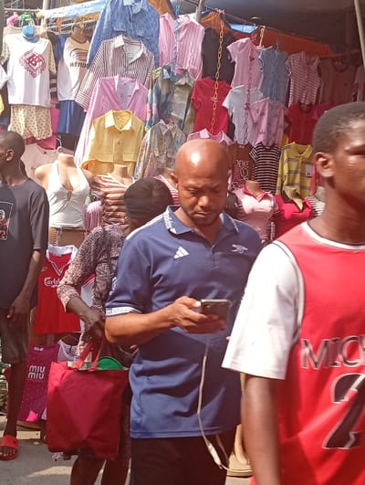 The image displays an outdoor clothing market in Asaba, Nigeria, characterized by bright daylight and pedestrian activity. In the center-foreground, a bald adult male in a navy blue Adidas polo shirt, featuring an Arsenal logo on the left chest, is focused on a black smartphone held in both hands, from which a white cable extends. To his right, a partially visible male wears a red sleeveless jersey with "MICHAEL 23" visible on the back. To his left, another partially visible male wears a dark shirt and red flip-flops. Behind the central male, a female carries a red fabric tote bag containing a green object. The background consists of a densely packed clothing stall, presenting numerous shirts, t-shirts (some with visible brands such as Burberry, WEIDONG, LOEWE, miu miu, and Chanel), and tops on torso mannequins. These garments hang from racks and ropes, with a partial blue tarp awning covering the stall overhead.