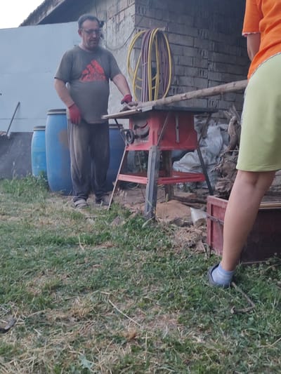 The image shows a man in a green t-shirt and dark pants, wearing red gloves and glasses. He is standing in what appears to be a yard or outdoor workspace, with a blue barrel and some machinery in the background. The setting suggests a rural or semi-rural location in Kochmar, Bulgaria. The man is holding onto a wooden plank, and his posture suggests he might be working on something or in the process of moving materials. There are also some coiled hoses in the background, which could be related to the machinery. The overall lighting suggests it is daytime, and the presence of grass indicates an outdoor environment. The image is oriented sideways.