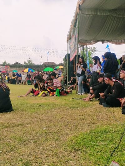 The image shows a gathering of people in an open-air space in Karangmalang, Indonesia (latitude: -7.47, longitude: 111.064). The scene appears to be a performance of traditional dance or theater. Several men, dressed in traditional costumes, are seated on the grass. The men are performing a dance or play, as indicated by their stylized gestures. A group of people are watching the performance, sitting on the grass or standing under a large white canopy tent. Several women are standing and watching, some holding smartphones or other devices. There are various colorful umbrellas in the background. The environment is characterized by grass, trees, and some buildings. The image is taken from a slightly elevated viewpoint, looking down on the scene.