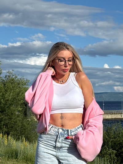 A young woman with blonde hair and glasses is posing outdoors. She is wearing a white crop top, a pink hooded sweatshirt draped over her shoulders, and light-wash jeans. A tattoo is visible on her stomach. The background features a bright blue sky with scattered clouds, lush green trees, and a body of water. The lighting suggests it is daytime, likely a sunny day. The overall mood appears relaxed and casual.