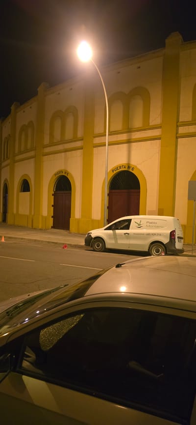 The image shows a street scene at night in Huesca, Spain. A yellow and white building with arched doorways labeled "PUERTA N° 5" and "PUERTA N° 6" is visible in the background. A white van with the name "Pablo Montero" and "Excavaciones - 685 909 216" is parked on the street. In the foreground, the hood and windshield of a silver car are visible, with reflections on the glass. An orange traffic cone is on the side of the road. There are no people visible in the image. The street appears to be paved, and there is a street light illuminating the scene.