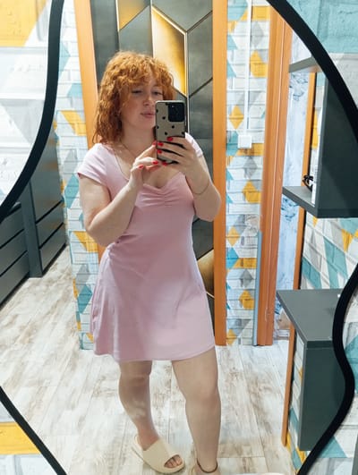 A red-haired woman in a pink dress takes a selfie in a mirror. She holds a phone with a black and beige dotted case and has red nail polish. She is wearing a gold bracelet on her left wrist and tan sandals. The background includes a wall with a pattern of geometric shapes in blues, yellows, and grays, wood trim, and a floor with wood-grain texture. A piece of black furniture is also visible. The location is Yurgamysh, Russia.