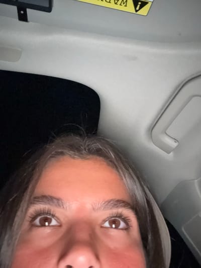 The image depicts a close-up, upward-angled view from the interior of a vehicle, primarily focusing on the upper portion of a person's face and the car's headliner. The specific location within West Windsor, United States, cannot be determined from the visual content provided.
**People:**
One individual is visible from the bridge of the nose upwards. The person has open brown eyes with prominent dark eyelashes and well-defined dark eyebrows. The skin on the forehead and upper cheeks appears a warm, possibly flushed, tone. Dark brown hair is visible, framing the forehead. The individual's gaze is directed slightly upwards, towards the camera or ceiling.
**Subjects/Objects:**
* **Vehicle Interior:** A light gray or beige fabric headliner with a textured pattern occupies the upper half of the image.
* **Sun Visor:** On the upper right side of the headliner, a portion of a yellow warning label, inverted, is visible, indicating its presence on a sun visor. Partial text, including "WARNING" and an exclamation mark, is discernible.
* **Grab Handle:** A light gray plastic grab handle is located on the right side of the headliner, partially visible.
* **Mounted Device:** In the upper left corner, a black rectangular object, possibly a rearview mirror casing or a dashcam, is mounted.
* **Exterior View:** To the left of the person's head and below the mounted device, a dark, undifferentiated area is visible, consistent with a vehicle window looking out into a nighttime or extremely low-light environment.
**Actions/Interactions:**
The person appears to be seated within the vehicle, looking upward. No specific action or interaction with other individuals or objects is discernible beyond the passive act of being present and observed.
**Environment:**
The scene is set inside a vehicle, likely in low-light conditions, as suggested by the dark exterior visible through the window. The primary light source appears to illuminate the person's face from below or the front.