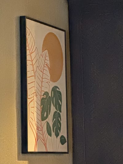 An framed abstract artwork featuring stylized green leaves and orange plant stalks against a pale background with a large orange circle representing the sun is leaning against a light tan textured wall. To the right of the artwork, a dark blue textured wall is visible, forming a corner with the tan wall. The scene is dimly lit, with a warm light casting shadows and highlighting the textures of the walls and the artwork. The image is taken from a slightly upward angle. There are no people or animals present. The location is not discernible from the image.