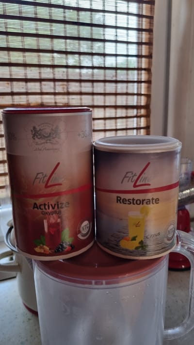 The image shows two cylindrical containers of "FitLine" products, likely dietary supplements, placed on top of a transparent pitcher. The container on the left is labeled "Activize Oxyplus" and features an image of a red beverage with berries and mint. The container on the right is labeled "Restorate" and depicts a yellow drink with lemon and mint. Both containers have the "FitLine" logo prominently displayed. The background consists of horizontal blinds, suggesting an indoor setting. The scene is set in Солнечная Поляна-3 Снт, Russia.