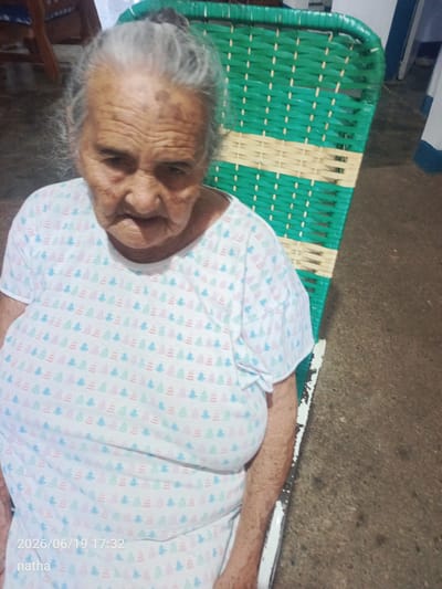 An elderly woman with gray hair is seated in a green and beige woven chair. She is wearing a white t-shirt with a pattern of small, colorful trees. The background appears to be an indoor setting with a concrete floor. A timestamp and the name "natha" are overlaid on the image, indicating it was taken on June 19, 2026, at 5:32 PM. The image was captured in El Chaparral, Venezuela.