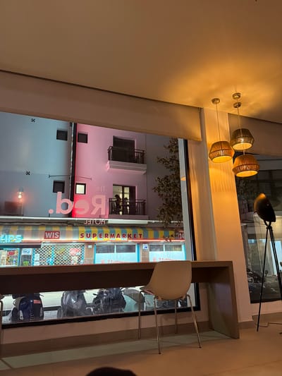 The image captures an interior view looking out through a large window onto a street scene at night in Sant Antoni De Portmany, Spain. The interior features a modern aesthetic with a minimalist desk and a white chair. Above the desk, three woven pendant lights cast a warm glow.

Outside, the street is illuminated by various signs and lights. A building with a pink facade is prominent, displaying a large, illuminated sign that reads "THE Red" in a stylized font. Below this, a store or supermarket sign is visible with "WIS SUPERMARKET" clearly legible, along with other signage that is partially obscured. The street level shows parked motorcycles and the reflective surfaces of shop windows. The overall impression is of a commercial street in a Mediterranean town at night, viewed from a comfortable interior space.