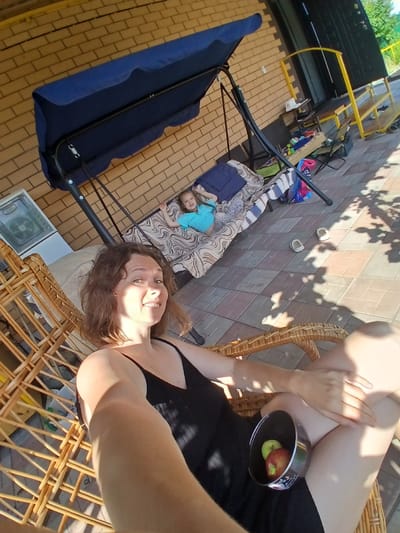 A woman sits in a wicker chair, holding a metal bowl containing two apples, one green and one red, in her lap. She is wearing a black dress. A young child is lying on a swing set with a blue canopy behind her. The scene is outdoors, with a brick wall and a paved patio visible. In the background, there is a yellow structure and greenery. The image appears to have been taken in Maslova Pristan', Russia, as indicated by the context.