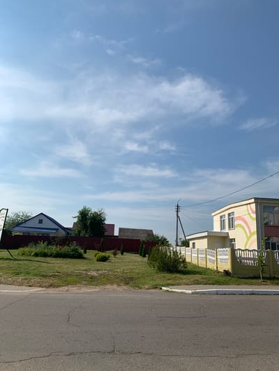 The image depicts a residential street scene under a bright blue sky with scattered white clouds, suggesting it is daytime. The orientation of the image is landscape, with the sky taking up the majority of the frame on the left, and buildings and a road on the right.
On the right side of the frame, a paved road runs horizontally, with a white curb separating it from a grassy area. Beyond the grass, there are several houses and fences. The first building visible is a multi-story structure with a pale yellow facade decorated with colorful, curved bands of pink, green, and yellow. It has multiple windows and balconies with white railings. Adjacent to this building is a yellow fence with decorative white elements. Further down the street, a dark red fence runs along the property line, behind which are more houses with dark roofs. One house has a prominent blue roof and white walls with a single small window visible.
A utility pole with wires is visible between the sky and the buildings. The overall atmosphere is tranquil and suggests a peaceful, suburban or rural setting. There is no visible text in the image. The time of day appears to be late morning or early afternoon, given the bright sunlight and clear sky. The weather is fair and sunny. The location is suggested to be Kapyĺ, Belarus. No people are visible in the image.