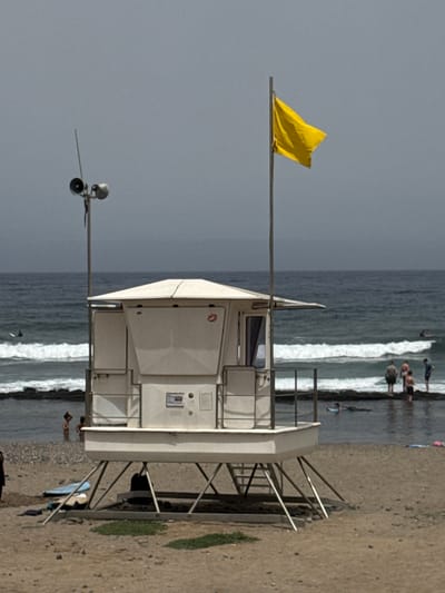 A bright yellow flag waves from a metal pole on a cloudy day at a beach. The flag is positioned above a white lifeguard tower. The tower, with its windows and railings, stands on a sandy beach next to the ocean. Gentle waves are breaking on the shore, where several people are visible, some standing in the water and others on the sand. The scene suggests a typical day at a coastal location, likely during daylight hours, given the presence of sunlight on the water and the visibility of the subjects. The sky is overcast, indicating a cloudy or possibly humid weather condition. The presence of the lifeguard tower suggests this is a monitored beach area, possibly a tourist destination, given the people enjoying the water. The overall atmosphere appears relaxed and typical of a beach environment. There is no visible text in the image.