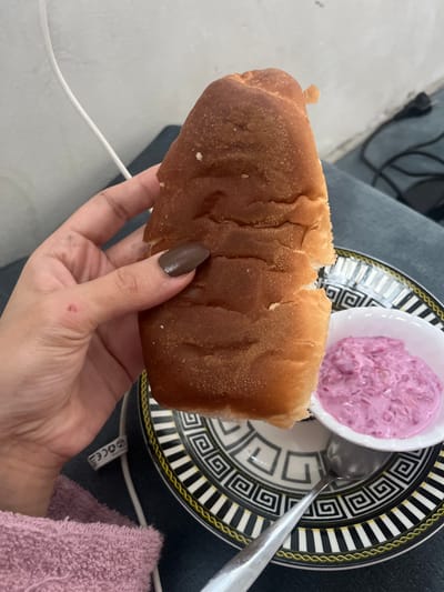 An indoor scene in Nuseirat, Palestinian Territory, captured from a first-person perspective, depicts a human hand holding a bread roll over a patterned plate containing a small bowl of pink dip.

In the foreground, a left hand with dark brown painted fingernails grips an elongated, golden-brown bread roll, which appears partially torn. A small reddish mark is visible on the back of the hand, near the base of the index finger. Below the hand, a fuzzy, light pink fabric is visible, likely a towel or blanket. A white charging cable runs across the lower left.

Centered in the middle ground, a round plate with a black and white Greek key pattern and a gold border holds a small, white, fluted ceramic bowl. The bowl contains a creamy, pinkish-purple mixture with visible textured elements, resembling a salad or dip. A silver-colored spoon rests on the plate next to the bowl.

The background shows a light-colored wall with some subtle texture. To the right, a dark blue or grey fabric, possibly clothing, is visible. An black electrical cord and a dark plug are also partially visible against the wall in the upper right quadrant of the image.