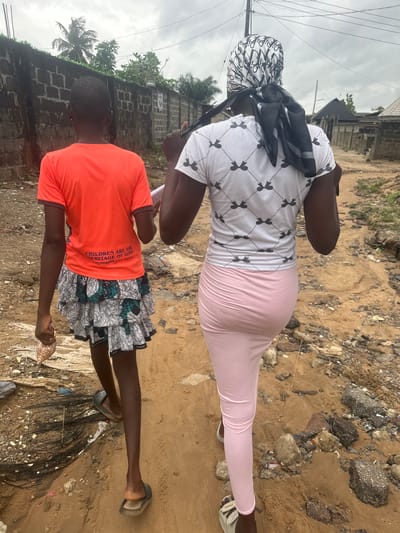 In the city of Ajara, Nigeria, two individuals, viewed from behind, are walking away from the camera on a dirt path. The person on the left is wearing an orange t-shirt with text that reads "CHILDREN ARE THE HERITAGE OF GOD Year 2022," and a patterned skirt. They are carrying a small plastic bag of what appears to be nuts in their left hand. The person on the right is wearing a white t-shirt with a repeating pattern of black bird-like figures and a light pink skirt or leggings. A black and white patterned scarf is tied around their head, with the ends hanging down. The environment is characterized by a rough, sandy terrain with scattered debris, including rocks and what looks like broken asphalt. A brick wall runs along the left side of the path, with trees and vegetation visible above it. A utility pole is present in the background. The sky is overcast.