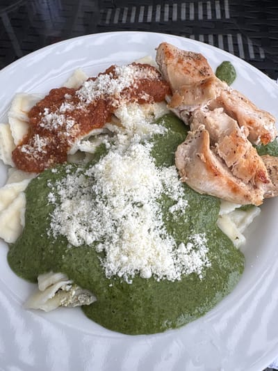 This is a close-up, eye-level, top-down shot of a white plate of food, likely taken at an outdoor dining establishment in San Antonio, Peru. The plate contains ravioli covered with two different sauces: a reddish-brown sauce and a bright green sauce, both topped with shredded white cheese. To the right of the ravioli are pieces of grilled chicken breast, also sprinkled with cheese. The background appears to be a dark, textured surface, possibly a table, and hints of a woven pattern suggest an outdoor setting. No people are visible in the frame.