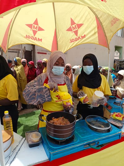 In the city of Tarauni, Nigeria, two women are preparing food under a yellow and red branded umbrella, possibly for a promotional event. The woman on the left, wearing a hijab and a blue surgical mask, is dressed in a patterned gray and blue abaya and a yellow apron with a Maggi logo. She is holding a yellow scoop with a Maggi cup, appearing to serve food from a large pot on a blue gas stove. The woman on the right, also wearing a hijab and a blue surgical mask, is in a bright yellow t-shirt and black pants. She is holding several white disposable cups and a yellow Maggi cup, seemingly ready to serve. Behind them, a crowd of women and girls, most wearing hijabs in various colors, are gathered, observing the food preparation. The setting appears to be an outdoor market or street stall, with a green artificial grass surface visible in the foreground and buildings in the background. A bottle of cooking oil and other containers are on a white table. The overall scene depicts a lively outdoor food preparation and distribution activity.