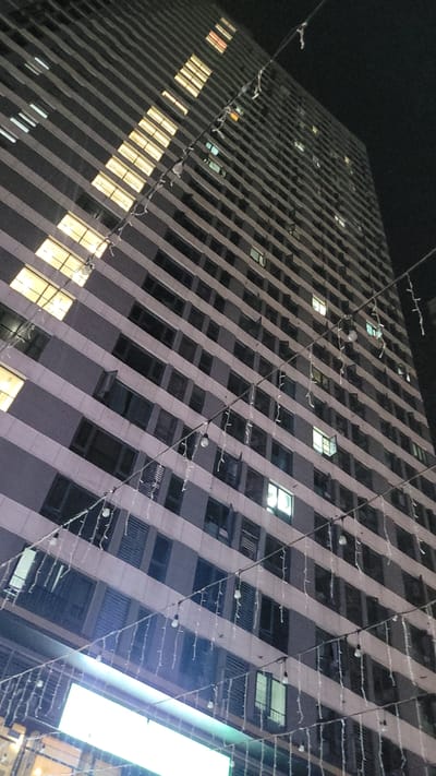 A low-angle view of a high-rise building at night, possibly in the Jiulongpo District of China. The building has a repeating pattern of windows across many floors, with some windows illuminated. String lights are draped across the building's facade. The sky is dark, suggesting it is night.