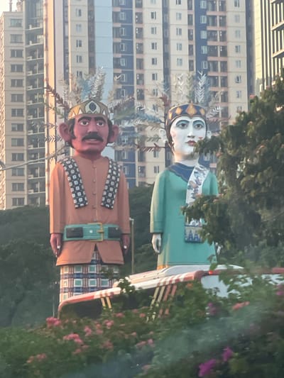 Two large, effigy-like figures are positioned in the foreground, in front of a backdrop of tall, modern buildings. The figure on the left has red skin, a mustache, and large ears, wearing an orange garment with a patterned scarf and a wide belt. The figure on the right has pale skin and is wearing a teal garment with a patterned sash. Both figures appear to be wearing ornate crowns. Behind them, several multi-story buildings with a grid-like pattern of windows dominate the scene. In the lower portion of the image, green foliage and pink flowers are visible, partially obscuring a vehicle with a red and white striped canopy. The lighting suggests it is daytime, possibly late afternoon given the warm tones on the buildings. This scene is located in Central Jakarta, Indonesia, where such figures, known as Ondel-ondel, are often displayed.