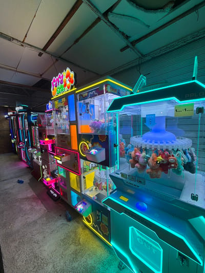 The image shows a row of brightly lit, colorful claw machines in what appears to be an arcade or entertainment venue. The machines are stacked and arranged in a line, each displaying a variety of plush toys within their transparent displays. Neon lights in various colors, including pink, yellow, blue, and green, illuminate the machines and the surrounding area, creating a vibrant and somewhat futuristic atmosphere. The floor is a rough, concrete surface, and the ceiling appears to be industrial, with visible pipes and corrugated metal. In the background, there are glimpses of more machines and a slatted metal door. One machine in the foreground has a sign that reads "LUCKY" and "taikongtu." The overall impression is that of a modern entertainment establishment, likely a place for games and prizes.

This scene is located at the GAMA amusement arcade in the city of Nesebar, Bulgaria.