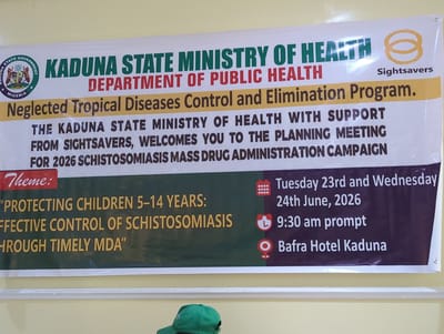 A banner hangs on a light-colored wall. The banner is predominantly white with sections of yellow, green, and dark purple. At the top left is the logo of Kaduna State Government with "Kaduna State Government Nigeria" written around it. Next to it, "KADUNA STATE MINISTRY OF HEALTH" is written in large green letters, followed by "DEPARTMENT OF PUBLIC HEALTH" in red. Below this is a yellow band with "Neglected Tropical Diseases Control and Elimination Program." written in brown. The main body of the banner contains text about a planning meeting. On the right side, a Sightsavers logo is present. Below the Sightsavers logo are details of the meeting: "Tuesday 23rd and Wednesday 24th June, 2026" and "9:30 am prompt" and "Bafra Hotel Kaduna." The theme of the meeting, in a green section on the left, is: "PROTECTING CHILDREN 5-14 YEARS: EFFECTIVE CONTROL OF SCHISTOSOMIASIS THROUGH TIMELY MDA". A person wearing a green cap is partially visible in the foreground, at the bottom of the image. The environment appears to be indoors, possibly a meeting room.
