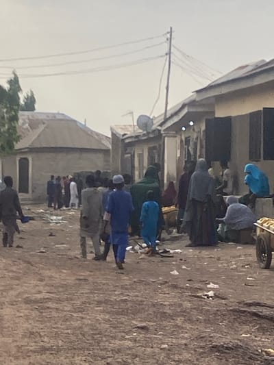 A street in Minna, Nigeria is depicted in this photograph. A group of people are walking along the street in both directions. Some individuals are wearing traditional clothing, including blue robes and caps. The street surface is littered with debris. Buildings line the street, with visible electrical wires above. A satellite dish is mounted on one of the buildings. The sky appears overcast.