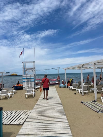 A bright, sunny day at a beach in Anapa, Russia. A wooden walkway leads across the sand towards the calm sea. A man in a red shirt and blue shorts walks away from the camera, carrying an orange bag. Several beach umbrellas and lounge chairs are set up on the sand. In the background, people are swimming and playing in the water. A yellow ferry is visible in the distance. A lifeguard tower stands near the water's edge, with a flag flying at the top. The sky is a vibrant blue, with scattered white clouds.