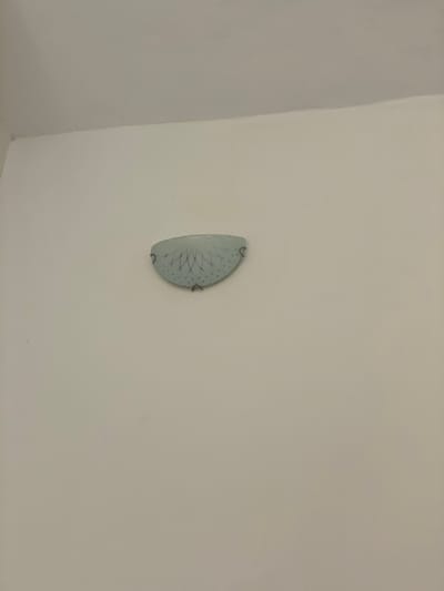 The image shows a white wall with a semi-circular object mounted on it. The object is light blue-gray and appears to have a decorative pattern of intersecting lines and small dots on its surface. The lighting is diffused, and the object is located approximately in the center of the frame, slightly to the left. The scene is indoors. There is no information available in the image or provided context to identify any details about people, subjects, actions, interactions, or the specific location in Ikeja, Nigeria.