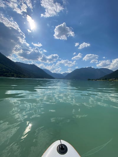 The image captures a serene scene on a lake with a stand-up paddleboard in the foreground on the right. The paddleboard is white with a textured surface and a black circular handle attached. The lake's water is a tranquil, greenish-blue, reflecting the sky and surrounding mountains. Ripples on the water's surface create patterns of light and shadow.

In the background, majestic mountains covered in dark green forests rise steeply from the lake's edge. Along the base of the mountains, a strip of green foliage hints at the shoreline, and a few buildings or structures are visible in the distance, suggesting a populated area. The sky above is a vibrant blue, dotted with fluffy white clouds. The sun is shining brightly, its rays breaking through the clouds, casting a bright glow.

The overall mood is peaceful and idyllic, evoking a sense of calm and enjoyment of nature. The scene appears to be set on a clear, sunny day, likely during daylight hours. The location is identified as Lärchenwiese, Austria. No text is visible in the image.