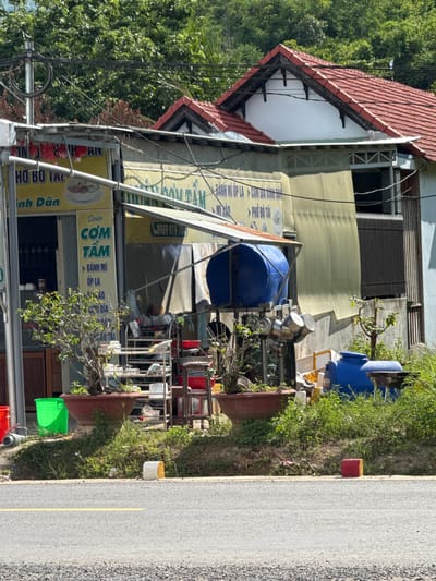 The image depicts a roadside scene in Nam Khanh Vinh, Vietnam, likely a small business establishment. In the foreground, a paved road with a yellow dividing line runs horizontally across the frame. To the right of the road, a low embankment is covered in green grass and vegetation. On this embankment, several objects are present, including a red cube, a white and yellow marker, and a small charcoal grill.
Behind the embankment, a modest building with a red tiled roof is visible. The building appears to be a shop or restaurant, with a yellow sign featuring Vietnamese text and images of food. Various items are displayed outside, including potted plants, shelves with kitchenware, large blue water tanks, and some red and green buckets. The area appears somewhat cluttered, with a mix of outdoor furniture and storage. The background consists of lush green trees and foliage, suggesting a semi-rural or suburban setting. The lighting indicates it is daytime, with bright sunlight casting shadows. There are no people visible in the image.
