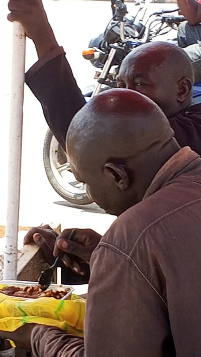 In Maiburiji, Nigeria, two dark-skinned men with shaved heads are seen in an outdoor setting. The man in the foreground is eating food, appearing to be beans, from a plastic container with a spoon. He is wearing a brown shirt. The other man stands behind him, with his left arm placed on a white pole. A motorcycle is visible in the background.