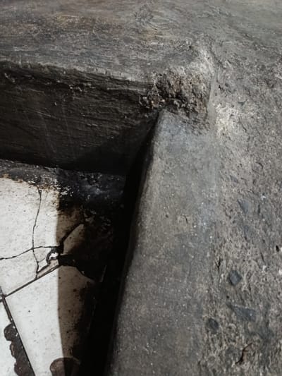 The image shows a close-up, high-angle shot of a weathered concrete structure adjacent to cracked white tiles. The concrete has a rough, uneven texture with visible aggregate. There are dark stains and patches on the tiles, suggesting spillage or wear. The lighting creates harsh shadows, emphasizing the textures and cracks. The scene appears to be an outdoor urban environment, possibly a neglected or aged part of a walkway or public space within Depok, Indonesia. No people or specific actions are depicted, only the static elements of the environment.