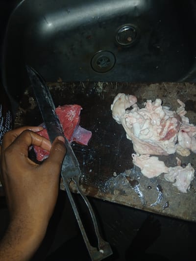 In Ikeja, Nigeria, a close-up shot captures a butcher's hands preparing raw meat in a dimly lit kitchen. The scene unfolds on a worn wooden cutting board, adjacent to a stainless steel sink. A person's left hand, with visible hair on the forearm, grips a large, sharp knife, poised to slice into a piece of red meat. The meat appears wet and glistens under the lighting. To the right of the red meat, a pile of white, fatty tissue and some red meat scraps are visible on the cutting board. The sink area, in the background, shows a drain and some water. The overall impression is one of a functional, albeit somewhat rustic, food preparation environment.
