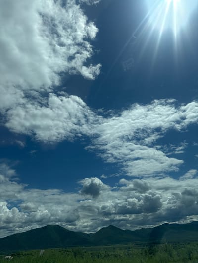 The image displays a wide shot of a mountainous landscape under a partly cloudy sky. The foreground is dominated by lush green vegetation, with a faint hint of a road or path visible in the lower left. In the mid-ground, a range of dark green, tree-covered mountains stretches across the horizon. The sky above is a vibrant blue, filled with a mix of white, fluffy clouds and some darker, more textured clouds towards the bottom. A bright sunbeam emanates from the upper right corner of the frame, casting rays across the sky and partially illuminating some of the clouds. There are no people or specific man-made structures clearly identifiable in the image, suggesting a natural, possibly rural or undeveloped area. The overall impression is one of a vast, open, and sunny day in a mountainous region.