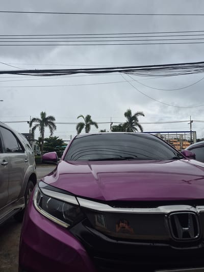 A purple Honda Civic is parked in front of a silver SUV on a cloudy day. The image is taken from a low angle, looking up at the cars and the sky. In the background, there are palm trees, buildings, and power lines. The license plate of the purple Honda is not visible. The scene appears to be in an urban or suburban area. The cars are wet, suggesting it may have recently rained. A reflection of a person taking the photo is visible in the grille of the purple Honda. The location is identified as Ban Yid, Thailand.