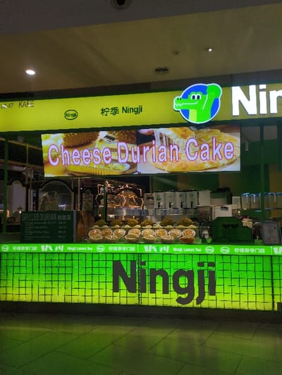 A brightly illuminated "Ningji" food stall is depicted within a commercial establishment in Sepang, Malaysia. The stall's upper fascia is primarily yellow, displaying the text "宁季 Ningji" alongside a green cartoon alligator head logo set within a blue circle. Below this, a large digital display showcases "Cheese Durian Cake" with associated photographic imagery of durian and a cheesy pastry.

The front of the service counter features a luminous green panel with a grid pattern, prominently displaying the "Ningji" brand name in dark lettering. On the counter, multiple rows of individually portioned, circular, yellow food items, possibly pastries or cakes, are presented in clear containers. Behind these items, several beverage dispensers and translucent storage containers are visible. To the left, a dark menu board lists "GRILLED DURIAN" offerings with prices denoted in Malaysian Ringgit (RM).

The immediate environment includes a dark, reflective floor surface and a light-colored ceiling with recessed lighting fixtures. No individuals are present in the foreground of the stall. The text "47 KAFE" is partially visible on the upper left portion of the stall's structure.