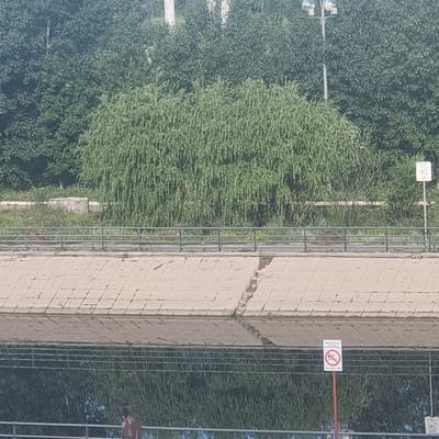 The image shows a concrete embankment with a railing along the top. Beyond the railing, there is a dense line of trees and foliage. A white sign with a red circle and a line through a swimmer icon, indicating "no swimming," is mounted on a red pole to the right of the embankment. Below this sign, there are smaller directional signs with arrows. The embankment appears to be part of a canal or river in the city of Aktobe, Kazakhstan. The water in the canal is visible at the bottom of the frame, reflecting the trees and sky. A person in a brown jacket is walking on the path behind the railing.