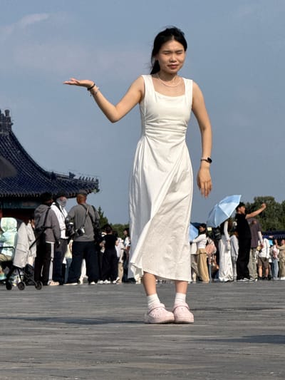A young woman in a white dress and pink Crocs stands on a wooden boardwalk, holding her left hand open, palm up, as if presenting something. She is in the foreground, facing slightly left, with her eyes squinted. Behind her, a crowd of people mills about. To the left, a traditional Chinese temple with a dark blue tiled roof is visible. The scene appears to be outdoors under a blue sky with some clouds. The woman is wearing a delicate pearl necklace and a pink fitness tracker on her left wrist. In the background, people are seen holding umbrellas, taking photos, and pushing a stroller. The location is Dongcheng District, China.