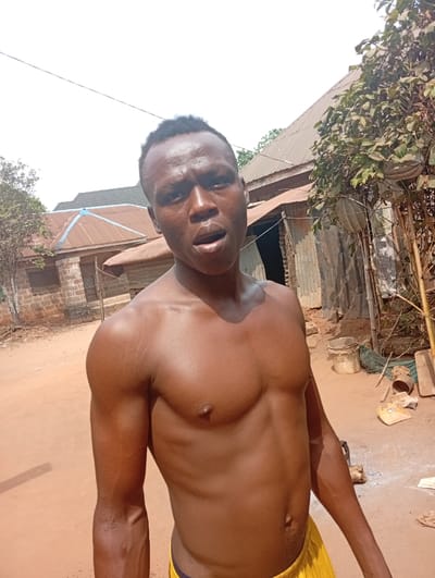 A shirtless man with dark skin stands outdoors. He has short, dark hair and is wearing yellow shorts. The man's body is muscular and toned. In the background, there are buildings with corrugated metal roofs, trees, and foliage. The ground is reddish-brown soil. The lighting is bright, casting shadows on the man's body. The overall scene appears to be in a rural or developing area.