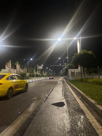 The image depicts a nighttime street scene in Jiulongpo District, China, characterized by a wet asphalt road surface suggesting recent precipitation. The environment is illuminated by numerous bright, horizontally-oriented streetlights mounted on tall poles, producing significant lens flares across the upper portion of the frame.

In the foreground to mid-ground on the left, a yellow sedan-style taxi, likely an Audi, is in motion, its wheels exhibiting motion blur as it travels away from the viewer. To the taxi's left, a blue and white segment barrier lines the roadside. Further along the left side of the road, a substantial, multi-sectioned retaining wall is visible. This wall features large, carved reliefs or murals, partially obscured by dark green foliage, possibly ivy or vines. Visible characters on the reliefs include "抗战精神" (Spirit of the War of Resistance) and "民族之魂" (Soul of the Nation), indicating a memorial or historical monument.

The road surface is dark asphalt, highly reflective due to moisture, and shows distinct white painted lane markings. A prominent, dark, roughly heart-shaped puddle is visible on the road surface in the mid-right.

On the right side of the road, a narrow grassy median with a concrete curb separates the main thoroughfare from a utility strip. This strip contains a light-colored utility box and a tall, slender pole. Scattered trees and bushes with dark green foliage are planted along this section. In the distance, further down the road, another dark-colored vehicle is visible, also traveling away from the camera. The sky above is dark and cloudless, contrasting with the bright artificial illumination. No individuals are explicitly visible in the frame.