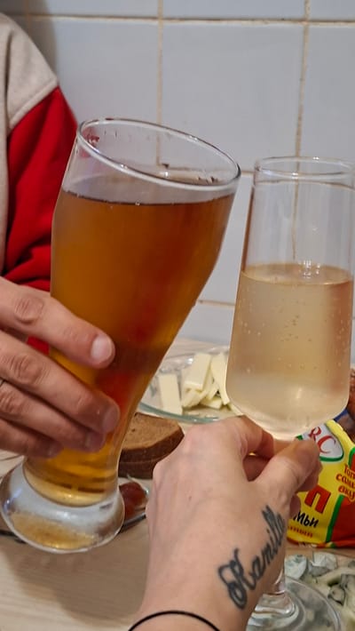 Two people are making a toast with drinks in a room with a tiled backsplash. On the left, a person in a red and tan jacket holds a tall glass of amber liquid, likely beer, with their left hand. On the right, another person holds a tall, clear glass of light-colored, bubbly liquid, possibly champagne or sparkling wine, with their right hand. Both hands are reaching across a table towards each other. In the background, there is a plate with slices of cheese and a piece of dark bread. To the far right, a bag with Cyrillic lettering and a yellow, green, and red design is partially visible. A tattoo of the name "Kamille" is visible on the right hand. The overall setting appears to be casual, possibly a meal or gathering in a home. The location is described as Забалоцкі сельскі Савет, Belarus.